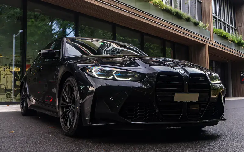 BMW M3 noire de trois quarts avant, large calandre M, garée devant un immeuble contemporain.