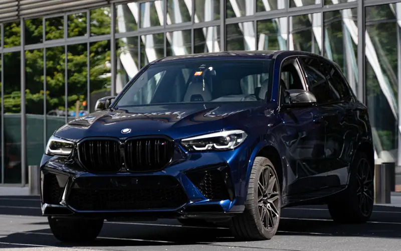 BMW X5 M Competition bleu Tanzanite vue de trois quarts avant devant une façade vitrée