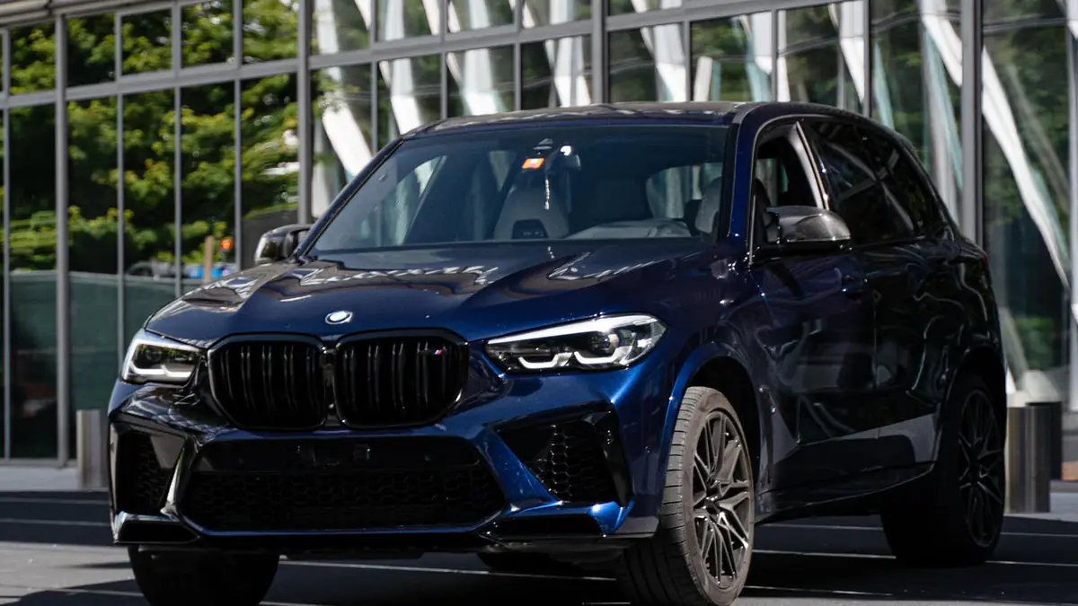BMW X5 M Competition bleu Tanzanite vue de trois quarts avant devant une façade vitrée