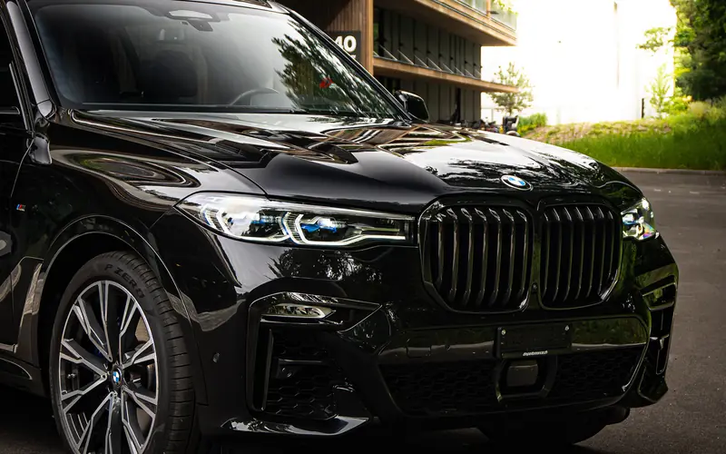BMW X7 M50i noir, vue de trois quarts avant, garé devant un immeuble en bois clair.