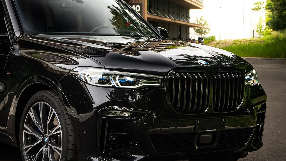 BMW X7 M50i noir, vue de trois quarts avant, garé devant un immeuble en bois clair.