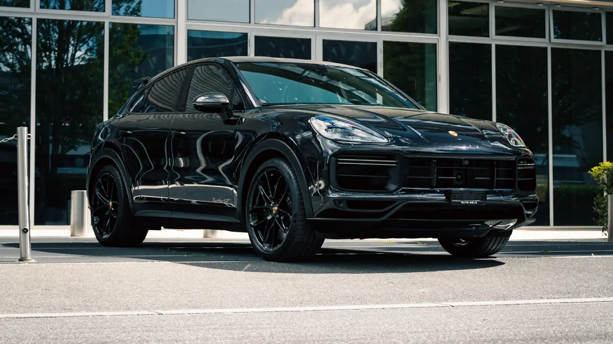 Porsche Cayenne Turbo GT noir vu de trois quarts avant, à l'arrêt devant une façade vitrée, en plein soleil.