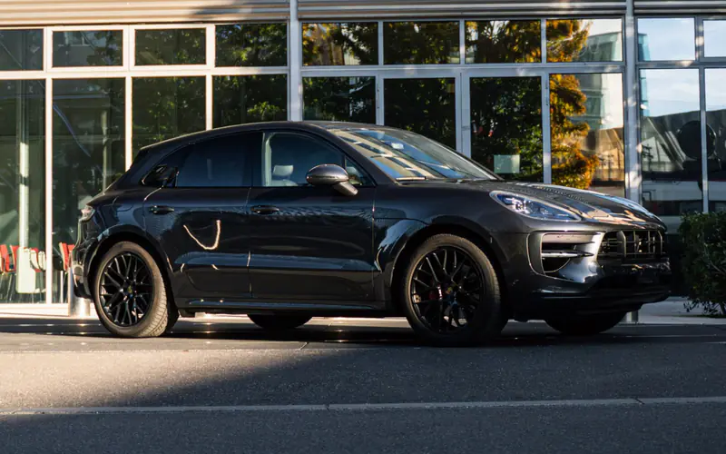Porsche Macan GTS noir vu de trois quarts avant, garé devant une façade vitrée en plein soleil.