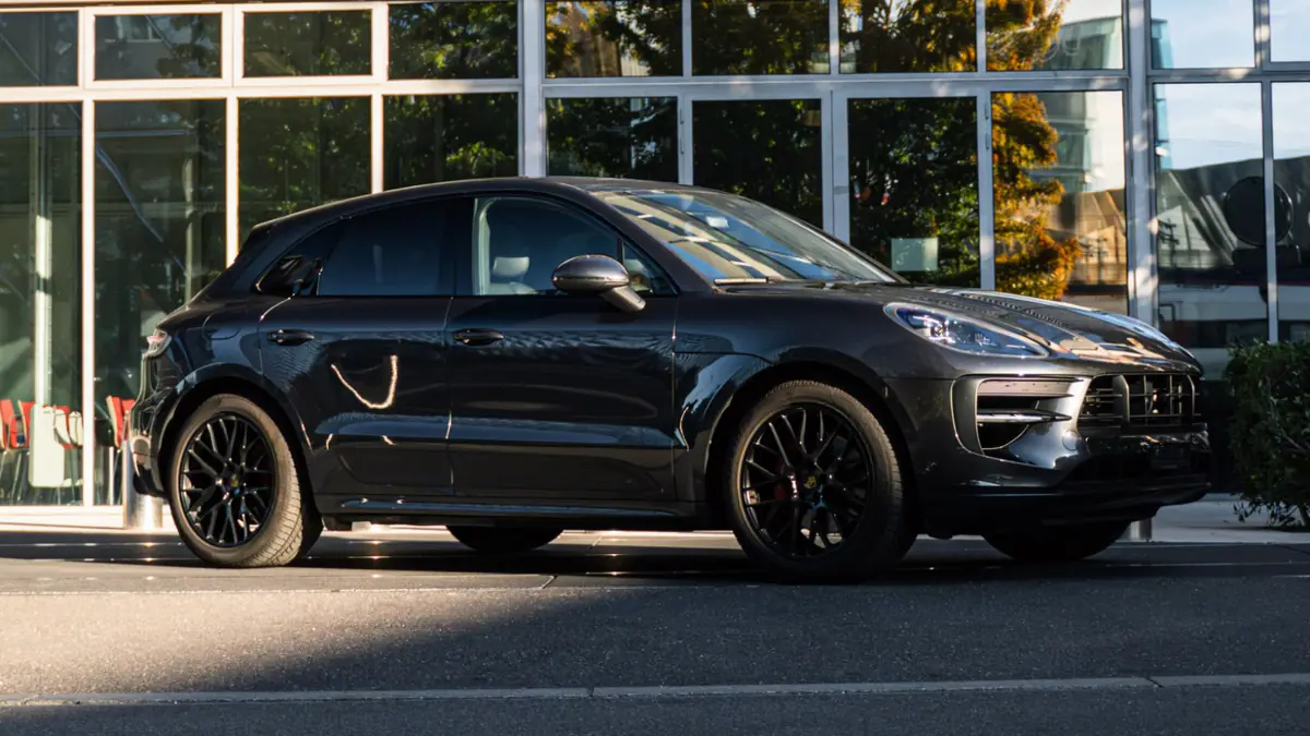 Porsche Macan GTS noir vu de trois quarts avant, garé devant une façade vitrée en plein soleil.