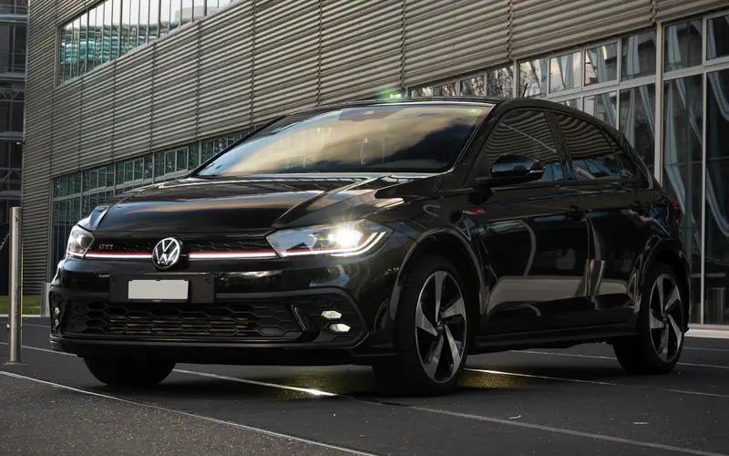Volkswagen Polo GTI noire vue de trois quarts avant, phares allumés, devant un bâtiment moderne.